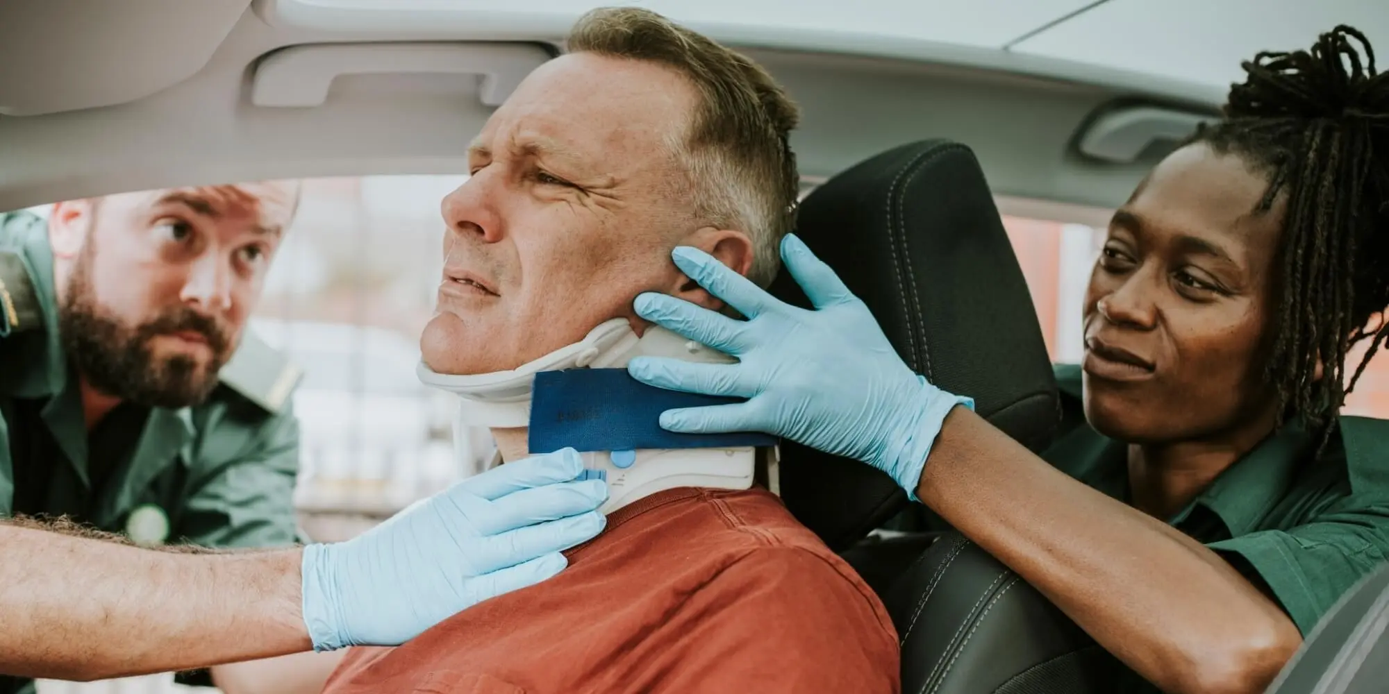Individual with a neck injury in a neck brace expressing pain, paramedics wearing medical gloves assisting with the individual's neck injury.