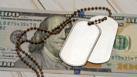 A $100 bill on a surface, silver military tags on top of the singular bill.